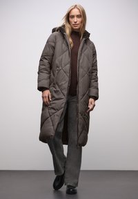 Long manteau matelassé rembourré de couleur gris foncé avec une capuche. Il présente des coutures diagonales, une fermeture éclair et des boutons-pression, et est assorti à un pantalon gris foncé.