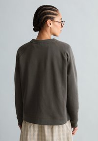 Person mit geflochtenen Haaren und Brille trägt einen dunkelgrauen Pullover und einen beigen karierten Rock, von hinten vor einem schlichten hellgrauen Hintergrund.