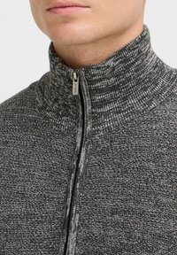 Chaqueta gris con cremallera y cuello alto, presenta un patrón de tejido texturizado y detalle de cremallera metálica. Tejido suave con un ajuste ceñido.