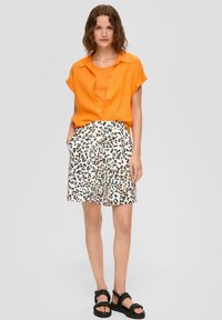 Pomarańczowa lniana koszula z guzikami w połączeniu z białymi krótkimi spodniami z czarno-pomarańczowym wzorem w lamparta. Czarne sandały na paski dopełniają całość.