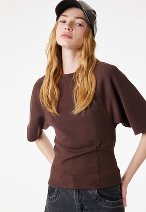 Giovane con capelli lunghi e ondulati, maglietta marrone a maniche corte, cappellino nero e jeans scuri, in posa con le mani sui fianchi contro uno sfondo bianco.