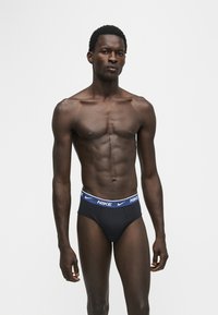 Homme en forme debout portant un slip noir Nike avec une ceinture bleue, face à un fond blanc uni.