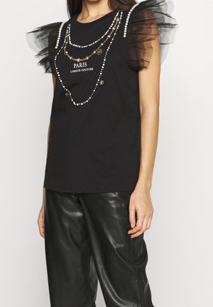 Nainen, jolla on musta hihaton paita, jossa on läpikuultavat rypytetyt olkapäät, helmien ja kultaketjun kuviointi, sekä mustat nahkahousut.