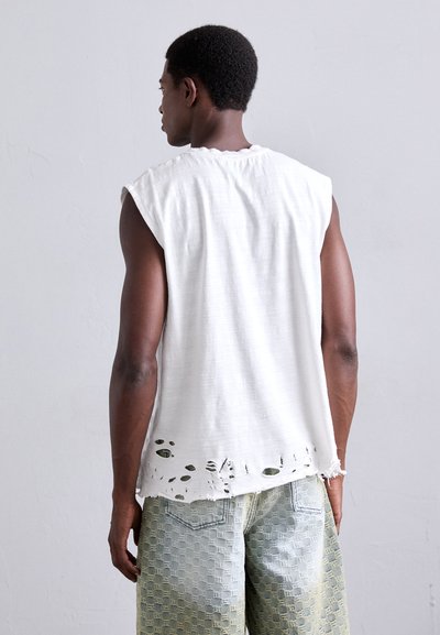 Hvid ærmeløs skjorte lavet af tekstureret stof med en revnet kant. Kombination med mønstret blå og grønne gradient jeans.