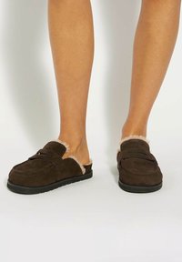 Brown suede clogs with a soft fur lining, slip-on design, rounded shape, and rubber sole. Features two crossed straps on top.