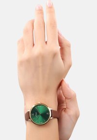 Runde Uhr mit grünem Farbverlauf-Dial, vergoldeter Lünette und großen Ziffern. Brauner Lederarmband. Hand auf dem Handgelenk ruhend, die Uhr zeigend.