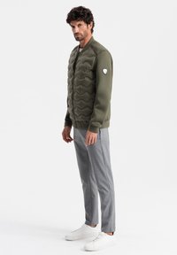 Olivgrön puffjacka med vågig textur, dragkedja och logotypdetalj på axeln, ihopsatt med ljusgrå byxor och vita sneakers.