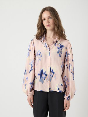 Kvinne med bølgete brunt hår iført en rosa bluse med blå abstrakte blomster mønstre og sorte bukser, stående mot en enkel lys bakgrunn.