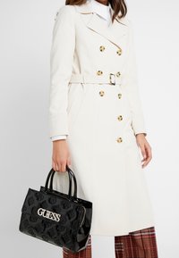 Trench-coat blanc croisé à double boutonnage avec boutons dorés et ceinture. Tient un sac à main en cuir verni noir avec logo embossé et motif floral.