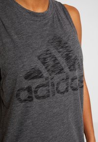 Šedý sportovní tílko s kulatým výstřihem, na přední straně velké černé logo Adidas a měkká, texturovaná tkanina.
