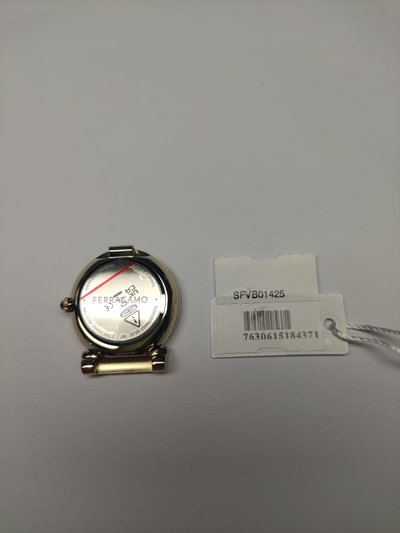 Kuldtooniga kell, millel on ringikujuline kellanupp, valge sihverplaat, punane tunniots ja brändi logo. Kaasas ripats, millel on nähtav baarikood ja SKU number.