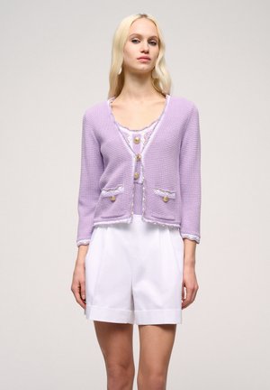 Cardigan tricotat lila cu un pattern texturat, cu fața deschisă și două buzunare; are accente contrastante deschise și butoane rotunde. Asortat cu pantaloni scurți albi.