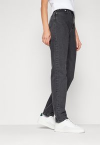 Jeans de mezclilla gris de pierna recta, con una cinturilla alta, un ligero desgaste, cinco bolsillos y remaches de metal. Combinados con zapatillas blancas.