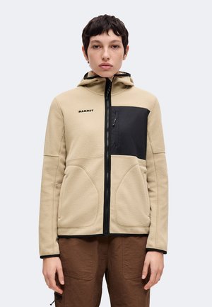 Persona con cabello corto y oscuro que lleva una chaqueta de forro polar beige con bolsillo negro en el pecho y pantalones marrones, de pie frente a un fondo claro y liso.