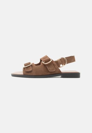 Καφέ σουέντ επίπεδο σανδάλι με μαύρη σόλα, ρυθμιζόμενα λουριά με βέλκρο και χρυσούς μεταλλικούς αγκράφες σε ουδέτερο φόντο.