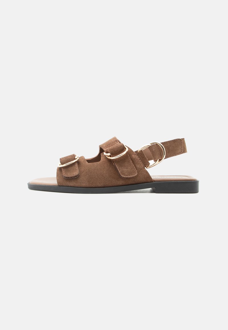 Καφέ σουέντ επίπεδο σανδάλι με μαύρη σόλα, ρυθμιζόμενα λουριά με βέλκρο και χρυσούς μεταλλικούς αγκράφες σε ουδέτερο φόντο.