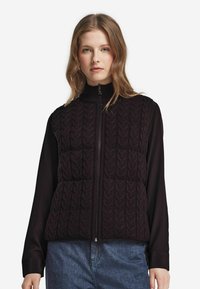 Ung kvinna iklädd en svart kabelstickad zip-up väst över en svart långärmad tröja och blå jeans, stående mot en enfärgad vit bakgrund.