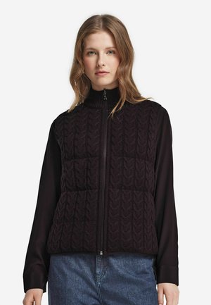 Junge Frau, die eine schwarze, kabelgestrickte Weste mit Reißverschluss über einem schwarzen Langarmshirt und blauen Jeans trägt, und vor einem schlichten weißen Hintergrund steht.