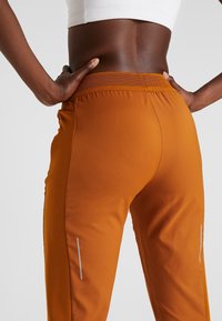 Oranger Sporthose mit einem enganliegenden Bund und reflektierenden Streifen an den Seiten. Glattes Material mit einer leicht dehnbaren Textur.