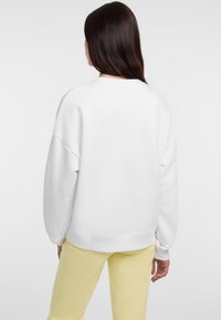 Sudadera blanca de gran tamaño con dobladillo redondeado y puños acanalados, combinada con pantalones amarillos claros, mostrada desde la parte de atrás.