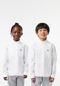 Due bambini indossano camicie bianche con bottoni e il logo Lacoste sul lato sinistro del petto, abbinate a pantaloni sportivi grigi. Tessuto morbido, maniche lunghe.