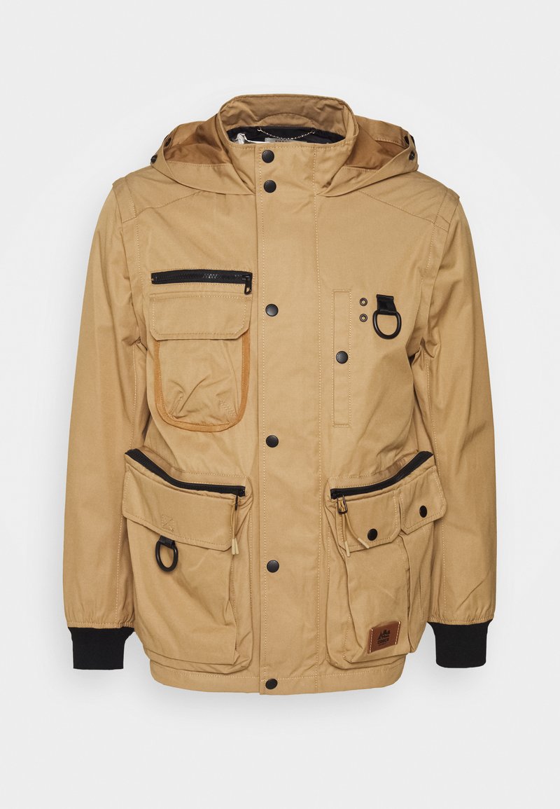 Veste beige avec capuche, plusieurs poches et accents noirs. Comprend des zippers, des boutons-pression et un tissu texturé pour la durabilité.