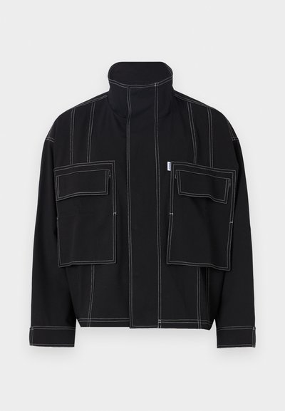 Black jacket made of cotton with high collar, featuring contrasting white stitching and two oversized front pockets. Simple, structured design.
