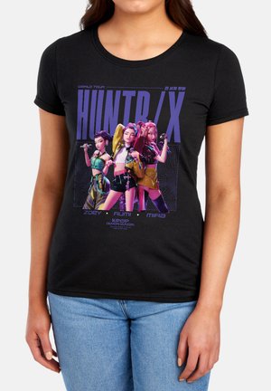 Three animated female characters posing with microphones under the text "HUNTR/X" on a black T-shirt worn by a person.
