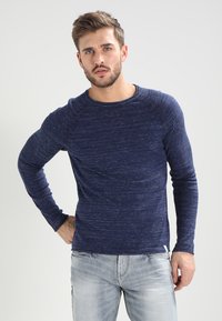 Camisa de manga larga de color azul marino con textura, que presenta un cuello redondo y acentos de canalé a lo largo de los hombros, combinada con unos vaqueros de denim claros.