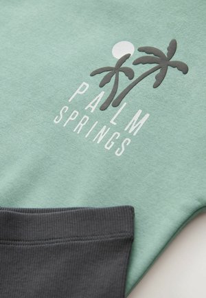 Mint green sweatshirt featuring “PALM SPRINGS” in white, with a graphic of two palm trees and a sun. Dark gray, ribbed cuffs. Soft texture.