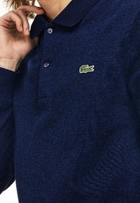 Námořnická modrá polo košile vyrobená z texturované látky s límečkem a dvěma knoflíky. Na levé straně hrudi je malé zeleně vyšívané logo.