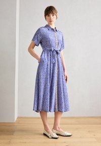 Vestido azul estampado com mangas curtas, um laço na cintura e uma frente com botões. Combinado com sapatos planos em creme com detalhes decorativos.