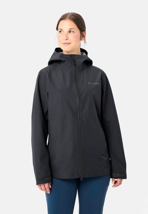 Vrouw met een zwarte waterdichte jas met capuchon en een ritssluiting aan de voorkant, en een blauwe broek, staand tegen een effen lichte achtergrond.