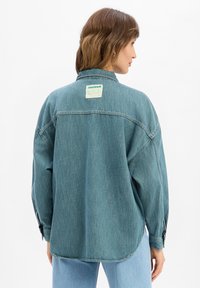 Veste en denim bleu clair surdimensionnée avec un ourlet arrondi, des poignets à boutons et une étiquette verte à l'arrière. Tissu doux et texturé visible.