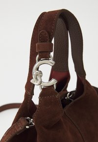 Geantă de mână din suede maro cu o curea din piele netedă, având un închizător argintiu și un interior roșu. Accentele includ cusături duble și un logo metalic.