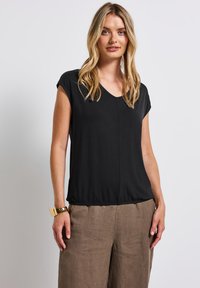 Svart kortärmad topp med V-ringning och något samlad nederkant, tillverkad av mjukt tyg, bärs med ljusbruna linbyxor.