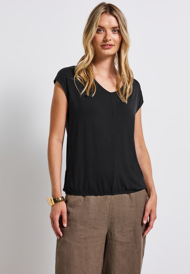 Svart kortärmad topp med V-ringning och något samlad nederkant, tillverkad av mjukt tyg, bärs med ljusbruna linbyxor.