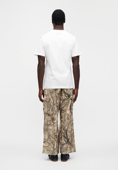 Λευκό βαμβακερό κοντομάνικο t-shirt, σε συνδυασμό μ ε μπεζ παντελόνι cargo με θήκες και σχέδιο καμουφλάζ δάσους, φορεμένο με μαύρα παπούτσια.