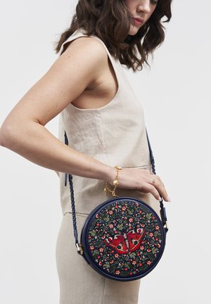 Femme portant une tenue beige sans manches tient un sac rond bleu marine avec un papillon rouge brodé et un motif floral, par une anse tressée.