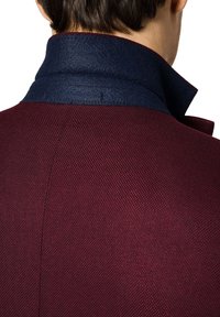 Burgundowa kurtka o teksturowanym splocie, z wysokim niebieskim filcowym kołnierzem. Widoczna staranna przeszycie wzdłuż krawędzi kołnierza.