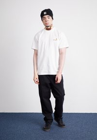 Maglietta bianca in cotone con un piccolo logo rosso e giallo, abbinata a pantaloni neri e sneakers nere, su uno sfondo semplice.