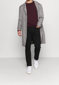 Man med en grå rutig lång kappa, vinröd tröja, svarta byxor och vita sneakers som går mot en enfärgad ljus bakgrund.