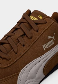 Brun ruskindsneaker med hvide og grå accenter, der har runde snørebånd, polstret tunge og gulddetaljer i logoet. Tekstureret materiale er synligt.