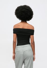 Kobieta o fryzurze afro, ubrana w czarną bluzkę na ramiączkach i szare spodnie w prążki, odwrócona plecami do jednolitego tła.