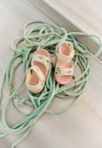 Ljusrosa sandaler med persika- och bladmotiv, tillverkade av flexibelt material, med justerbara remmar, placerade på grönt rep.