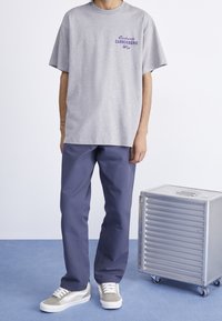 Person iført en grå t-shirt med lilla tekst, mørkeblå bukser og beige sneakers, stående ved siden af en metalopbevaringskasse med hjul.