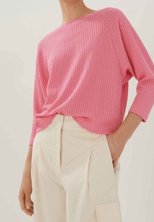 Femme portant un haut rose texturé à manches longues rentré dans un pantalon plissé taille haute de couleur blanc cassé, une main dans la poche.
