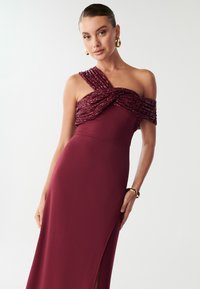 Burgundijska večernja haljina s jednim ramenom, koja se odlikuje uvijenim, teksturiranim korzetom i visokim prorezom do tableta. Glatka tkanina i elegantan kroj.