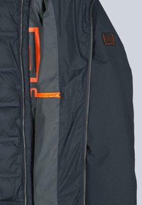 Innenansicht einer marineblauen Steppjacke mit grauem Futter, einer orangefarbenen Reißverschlusstasche und einem Logo-Patch in der oberen Ecke.