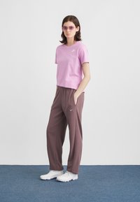 Νεαρή γυναίκα φοράει ένα ροζ μπλουζάκι Nike, καφέ φόρμες Nike, λευκά αθλητικά παπούτσια και γυαλιά ηλίου με μωβ απόχρωση, στέκεται σε ένα μπλε χαλί.